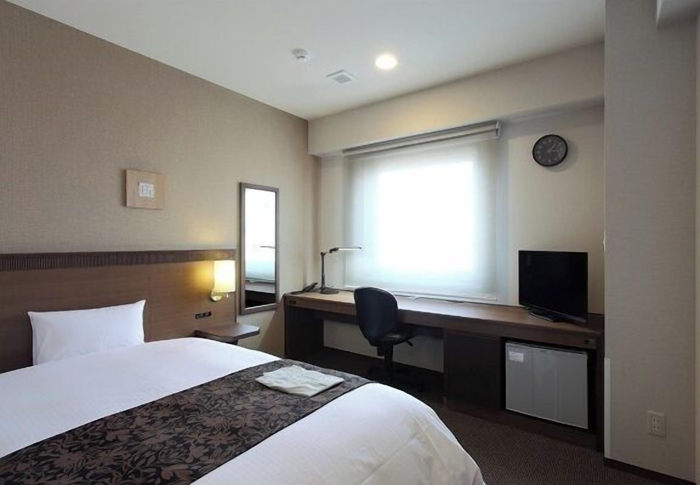 Habitación de hotel con cama, escritorio, silla, televisor y un reloj en la pared.