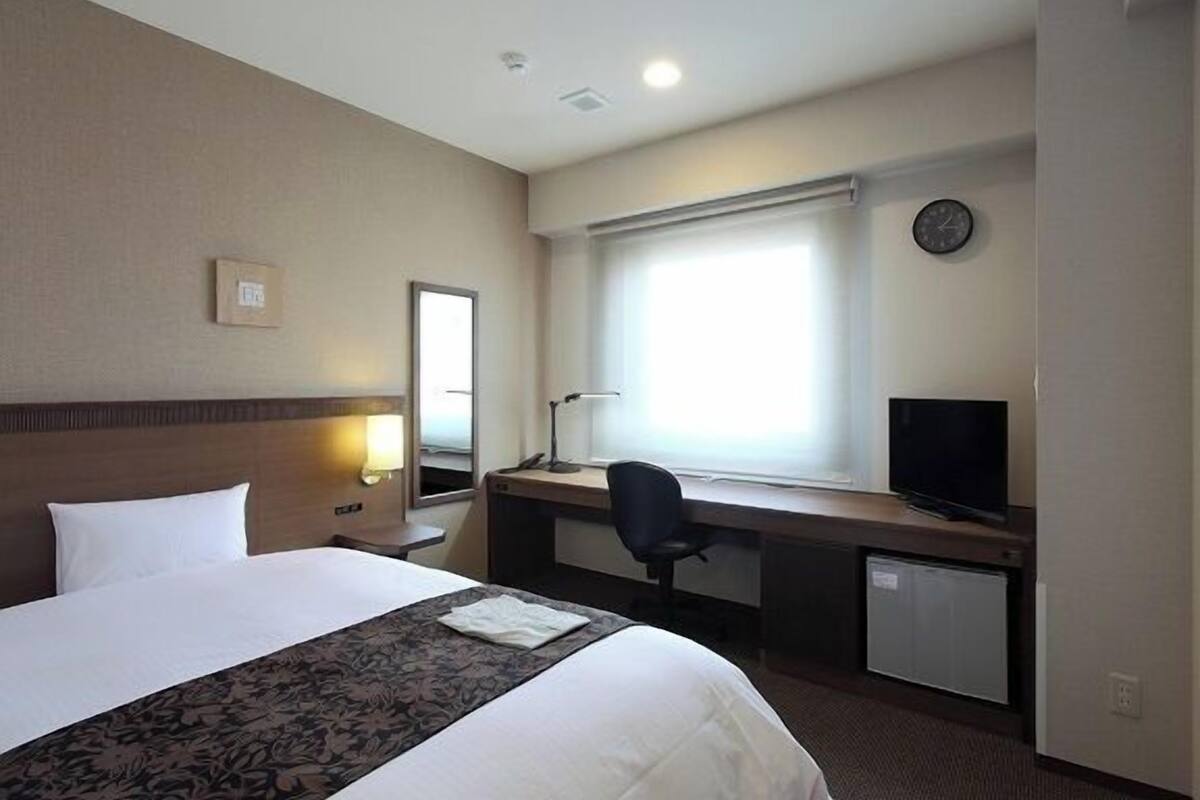 A hotel room with a bed, desk, chair, television, and a clock on the wall.