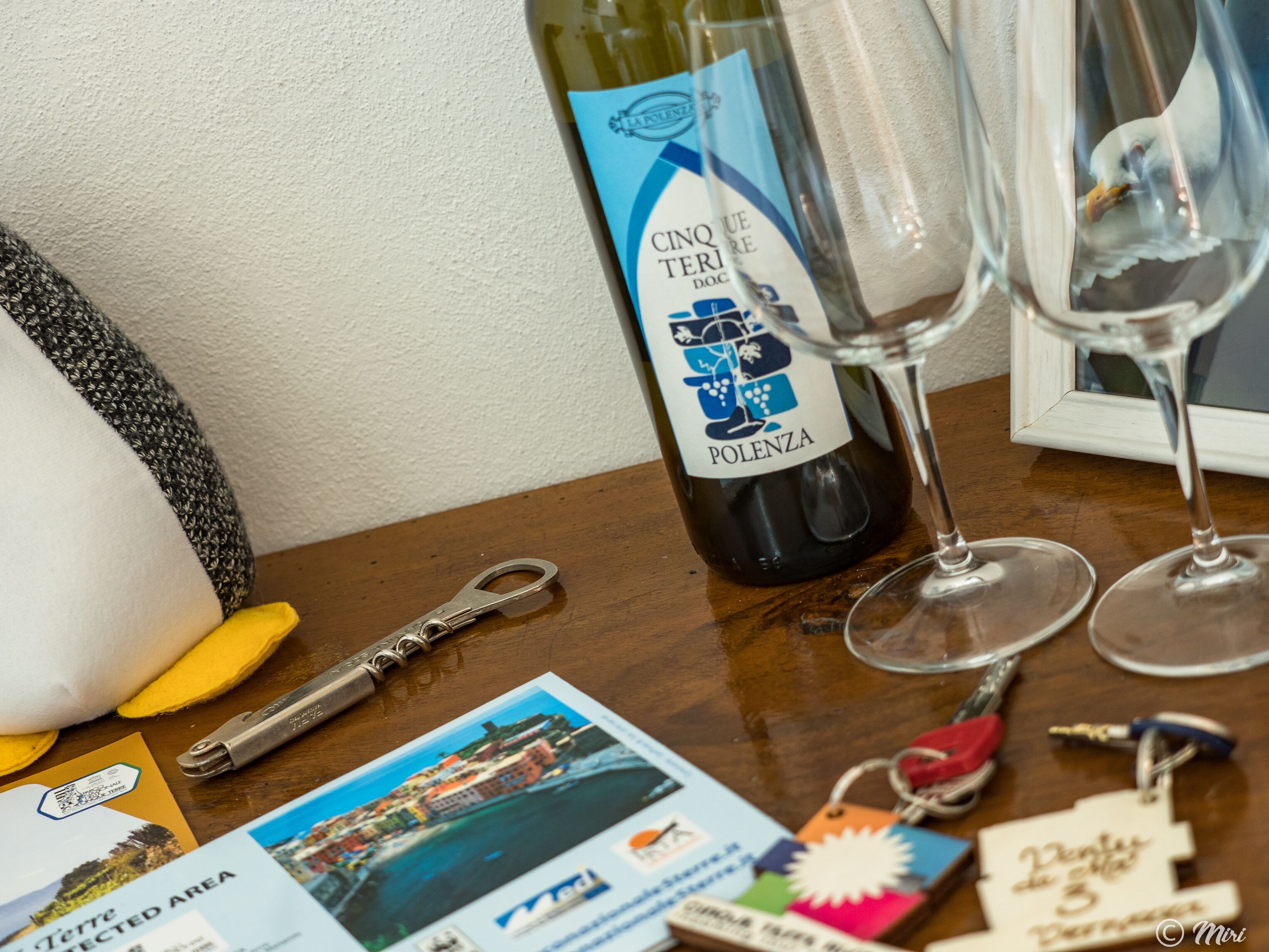 A bottle of Cinque Terre DOC wine, a wine glass, a key, a postcard, and a keychain on a wooden table.