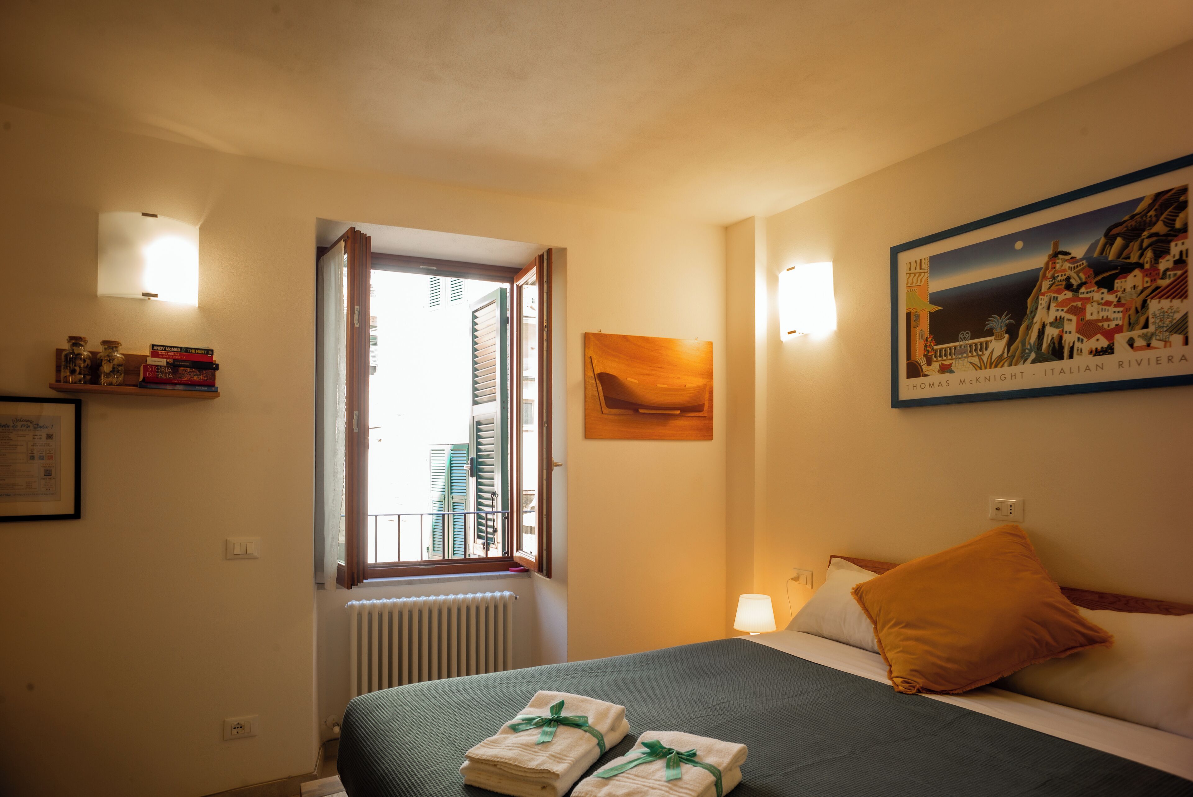 A hotel room with a bed, a window with shutters, a painting, and a lamp.