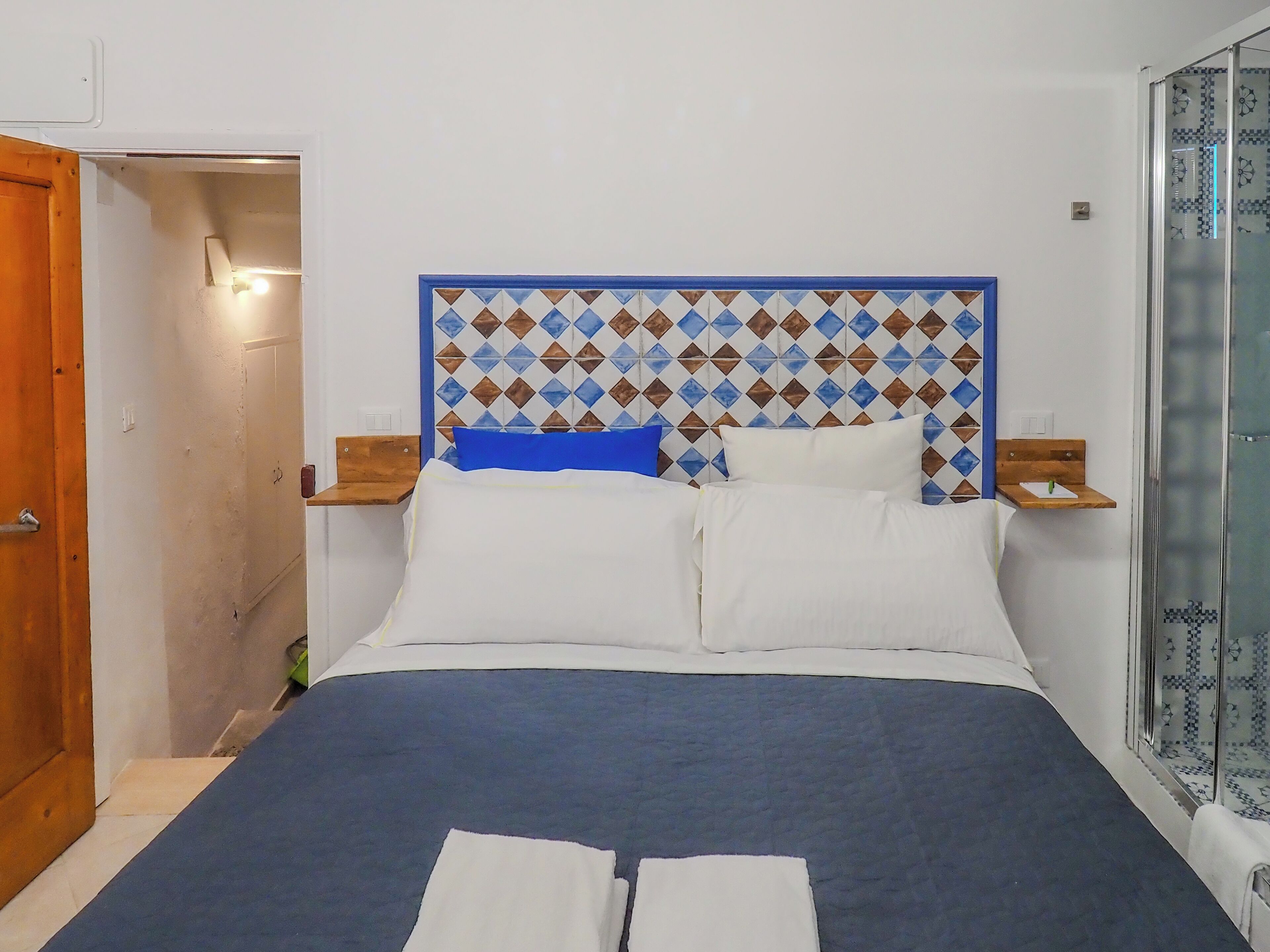 A neatly made bed with a patterned headboard, a bedside table with a lamp, and a glass shower enclosure.
