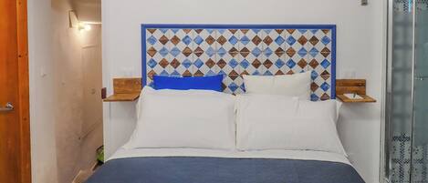 A neatly made bed with a patterned headboard, a bedside table with a lamp, and a glass shower enclosure.