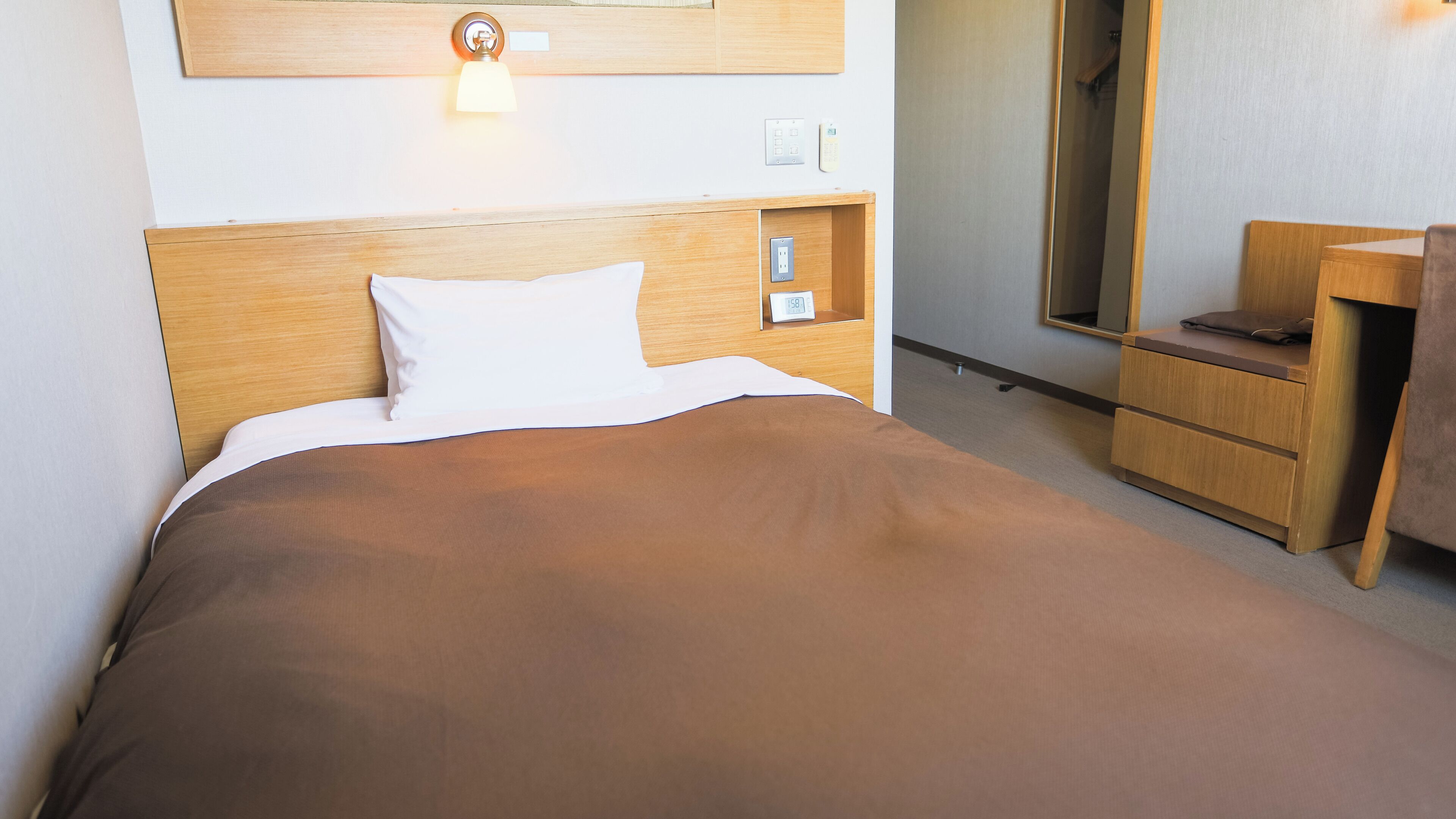 A hotel room with a wooden headboard, a bed with white and brown bedding, a bedside table with a lamp, a small clock, and a mirror on the wall.