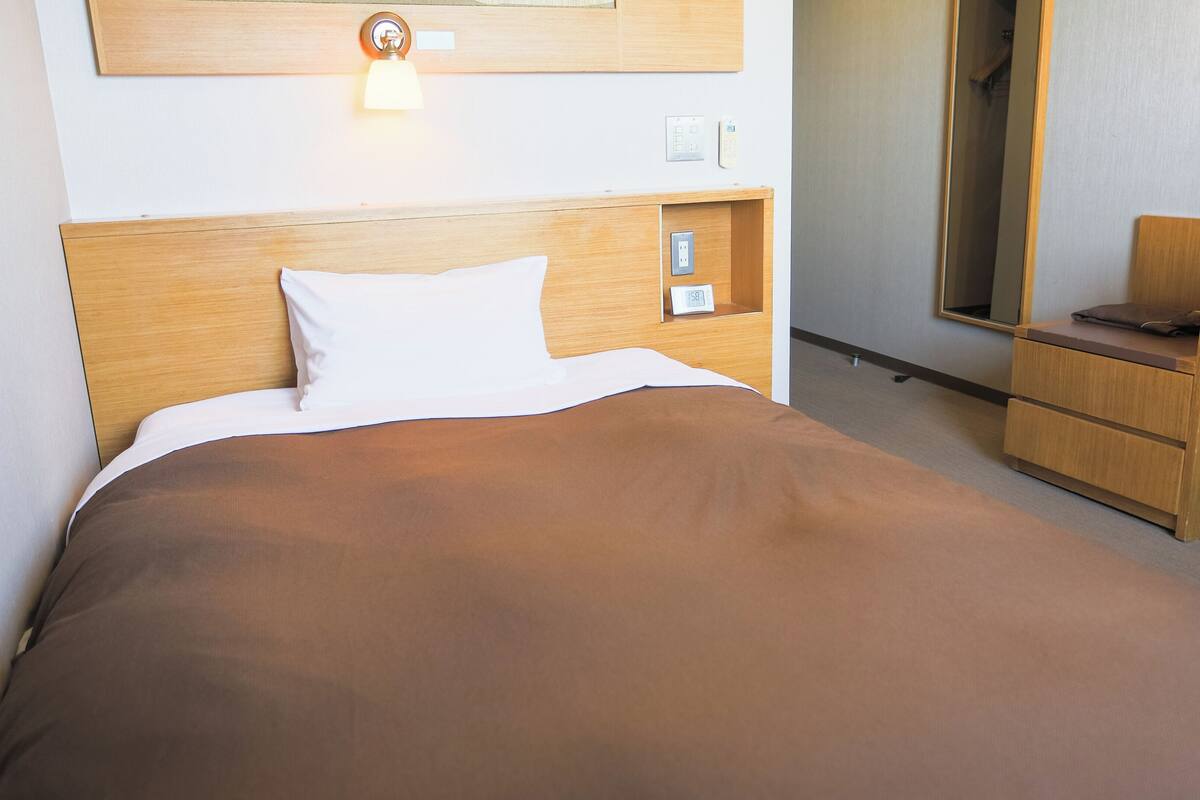 Hotellihuone, jossa on puinen sängynpääty, valkoisilla ja ruskeilla tekstiileillä varustettu sänky, yöpöytä lampun, pienen kellon ja seinällä olevan peilin kanssa.