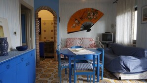 A room with a blue dining table, blue chairs, a blue sofa, a television, and a decorative fan. - Casa Ostrica (Alghero)
