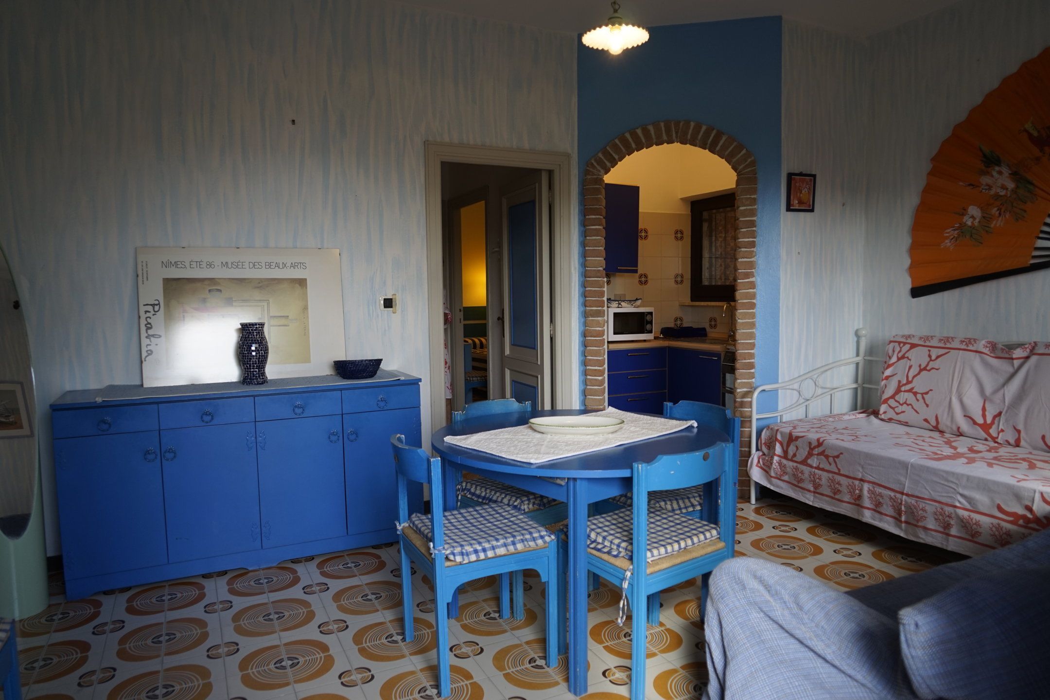A room with a blue cabinet, a dining table with chairs, a bed with a patterned bedspread, and a wall-mounted fan.