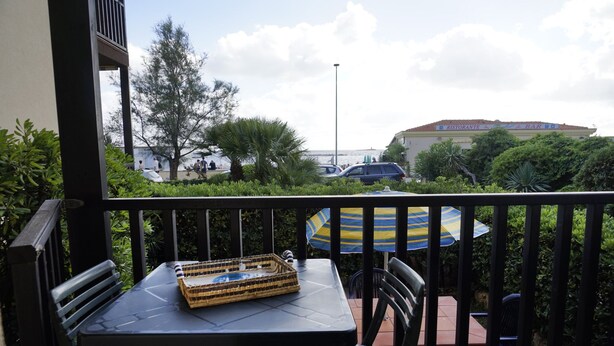 A balcony with a table and chairs, a striped umbrella, and a view of the sea and buildings in the background. - Casa Ostrica (Alghero)
