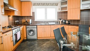 A modern kitchen with wooden cabinets, a stainless steel range hood, a built-in washing machine, and a glass dining table with chairs. - Beach Boulevard Apartment (Aberdeen)