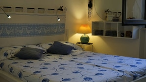 Una cama bien hecha con colcha floral, una lámpara de noche y un estante con diversos objetos.