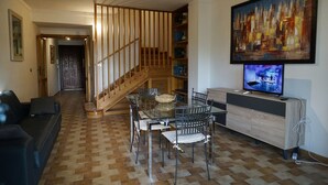 Una sala de estar con escalera de madera, una mesa de comedor con sillas, un sofá y un televisor sobre un mueble de madera.