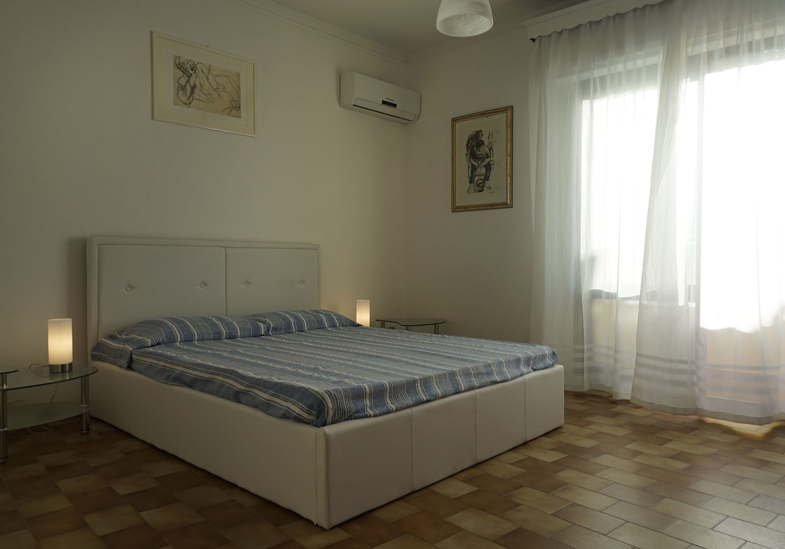 A bedroom with a large bed, two bedside tables, a wall-mounted air conditioner, and two framed artworks.