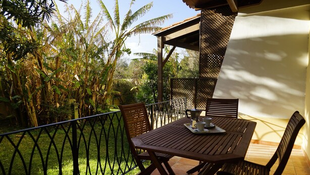 Un patio al aire libre con una mesa y sillas de madera, rodeado por una valla de metal y abundante vegetación.