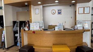 A reception desk with a wooden counter, a clock, framed certificates, and informational brochures. There are two black stools and a wooden bench in front. - HOTEL CROWN HILLS TSUCHIURAEKIHIGASHI (Tsuchiura)