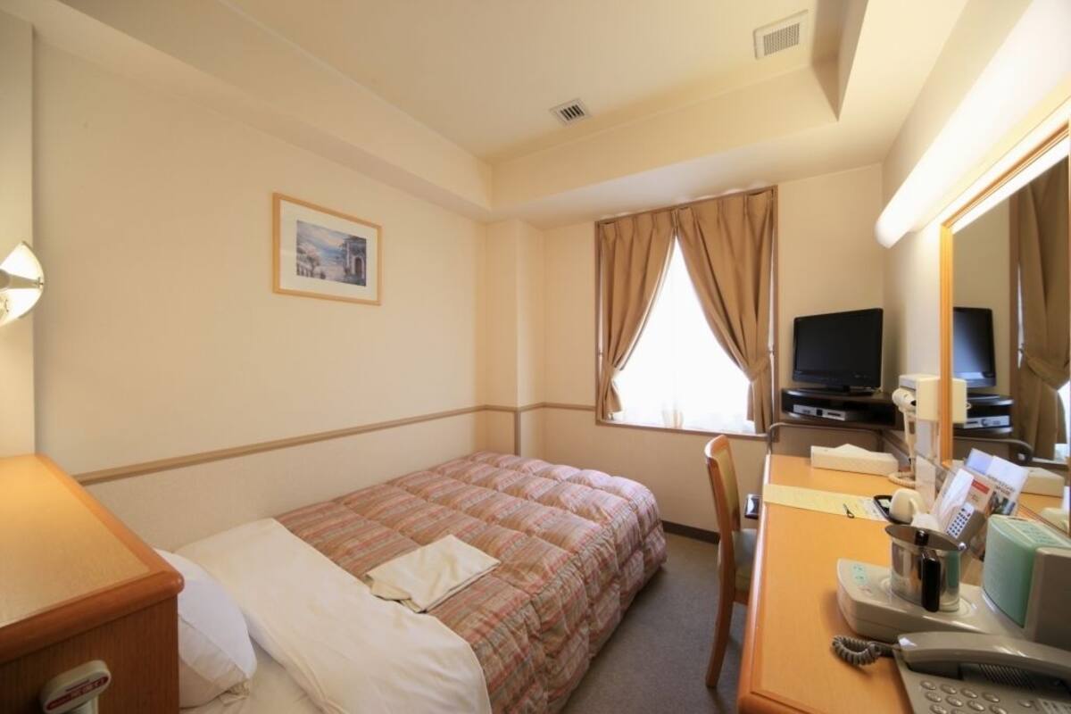 A hotel room with a single bed, a desk with a telephone, a television, and a window with curtains.