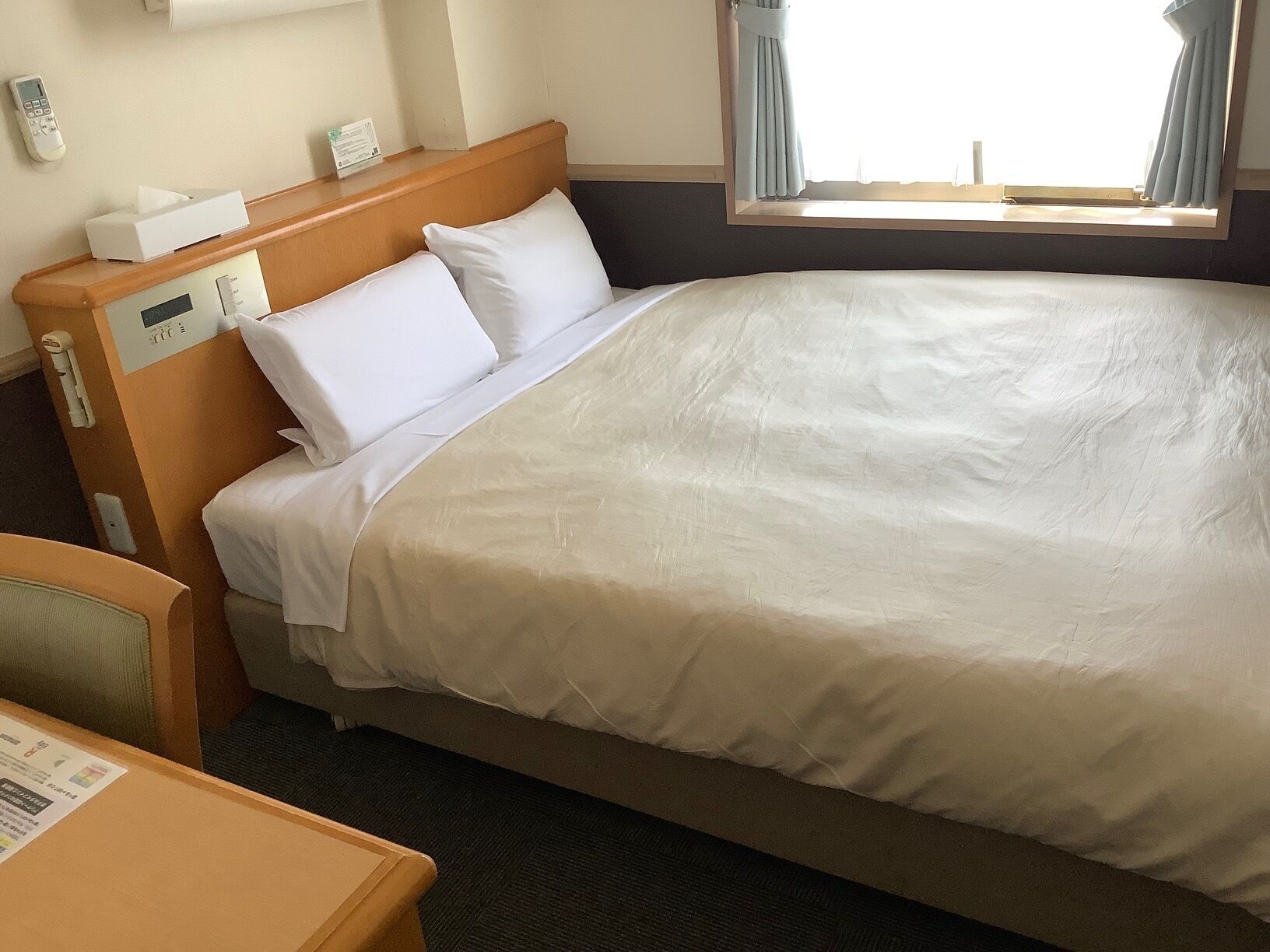 A hotel room with a large bed, a bedside table, a control panel, and a window with curtains.
