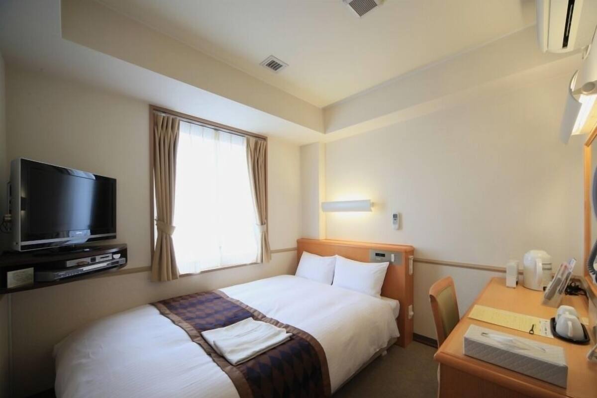 A hotel room with a bed, a TV, a desk with a coffee maker, and a window with curtains.