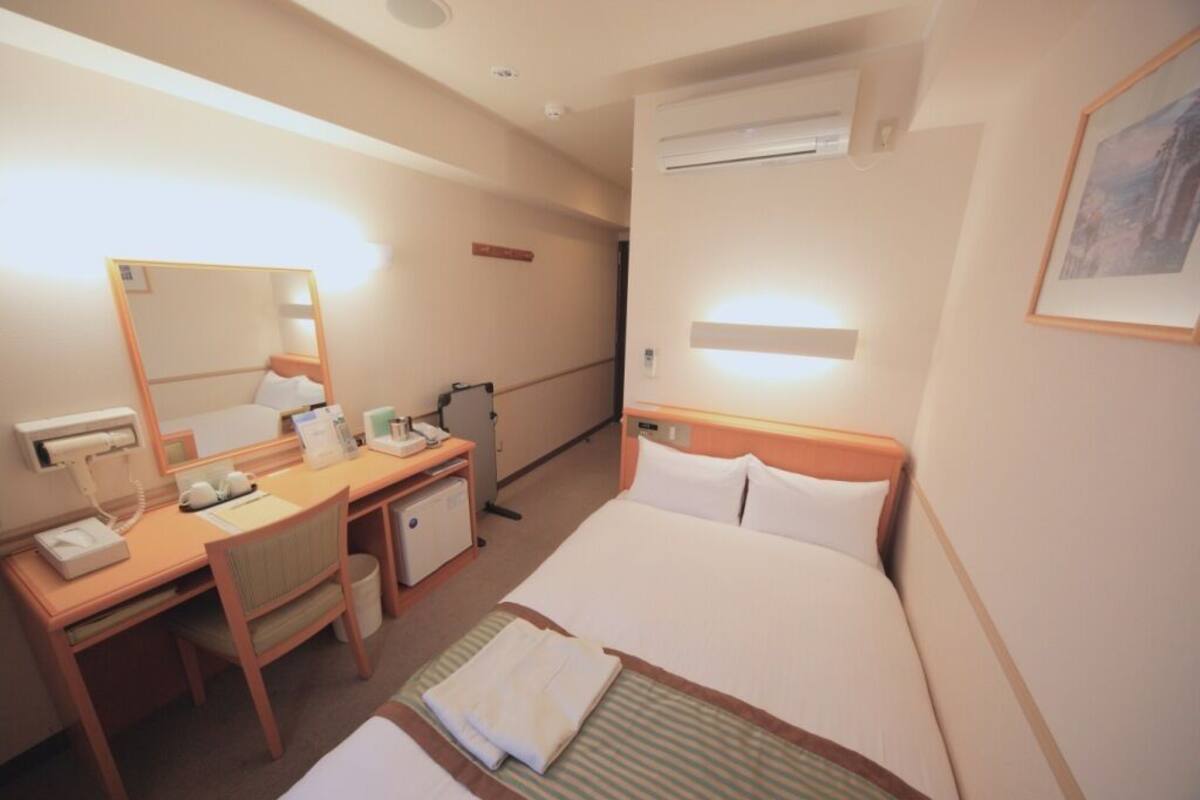 A hotel room with a bed, a desk, a chair, a mirror, a bedside table, and an air conditioning unit.