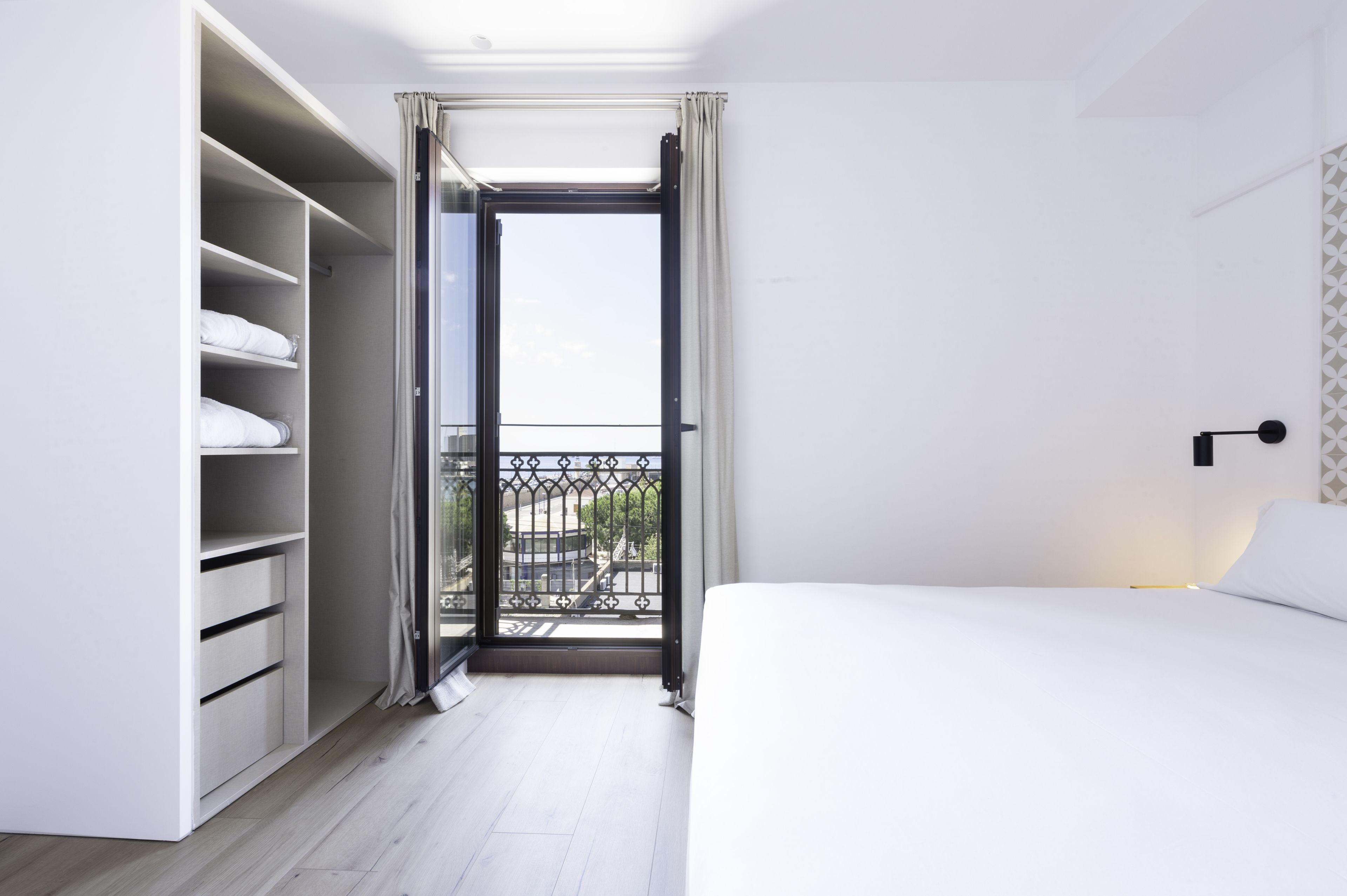 Ein modernes Hotelzimmer mit Balkon, einem weißen Bett und einem Kleiderschrank mit vielen Ablagen.