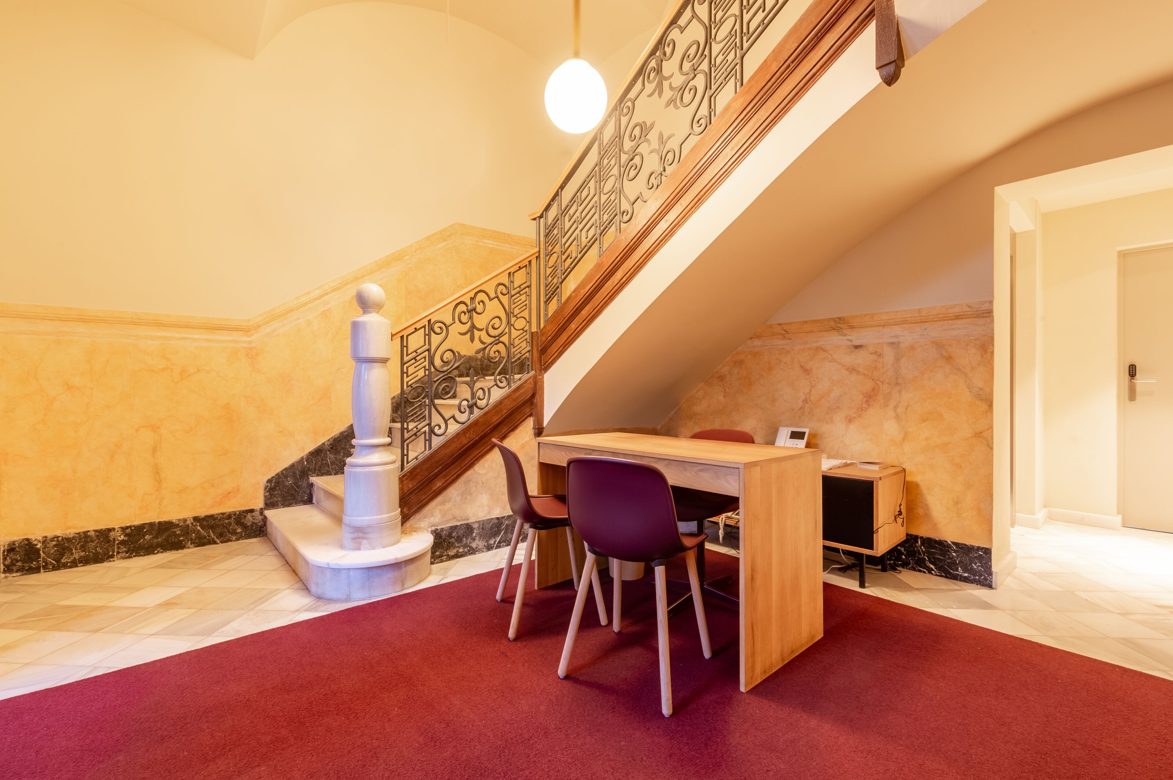 A room with a wooden desk, purple chairs, a marble wall, and a staircase with a decorative railing.