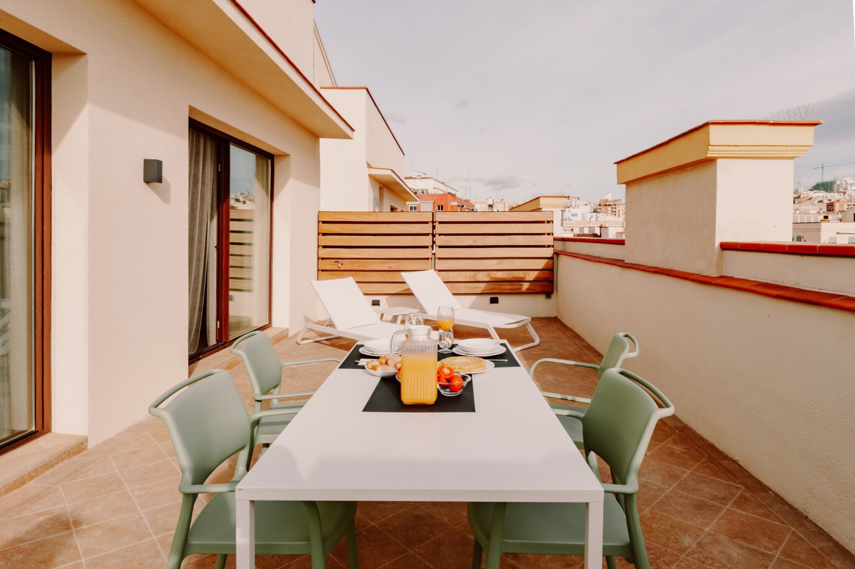 A rooftop terrace with a table set for breakfast, including a jug of orange juice, plates, and a bowl of fruit.