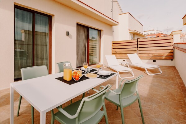 Un coin repas avec une table dressée pour deux personnes, une bouteille de jus d’orange et une assiette de nourriture. Il y a deux chaises et une chaise longue.