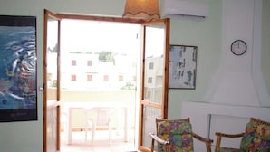 An open sliding door leading to a balcony with a table and chairs, a wicker chair with a floral pattern, and a wall clock. - Casa Calamaro (Alghero)