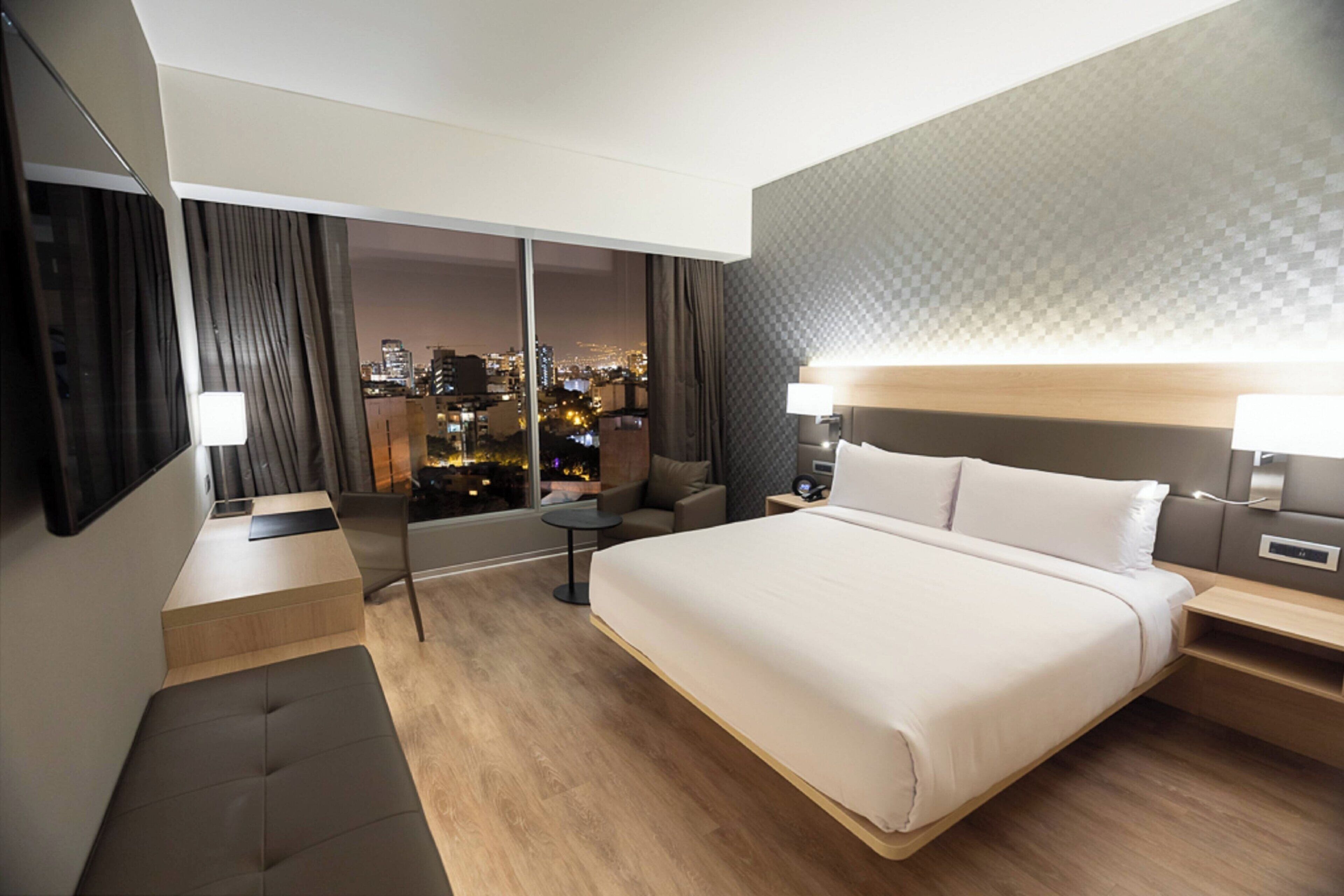 Een moderne hotelkamer met een groot bed, een bank, een bureau en uitzicht op de stad 's nachts.