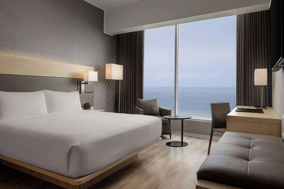 Une chambre d’hôtel moderne dotée d’un grand lit, d’un bureau avec une chaise, d’une petite table et offrant une vue sur l’océan grâce à de grandes fenêtres.