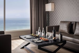 A modern hotel room with a leather sofa, a dining table set with plates, glasses, and bottles, and a view of the ocean.