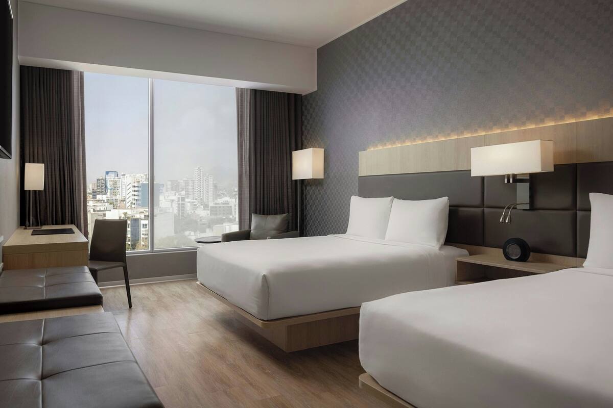 Une chambre d’hôtel moderne avec deux lits, un bureau et une grande fenêtre donnant sur la ville.