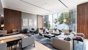 Un moderno hall de hotel con diversas zonas de estar, incluyendo sofás y sillones, organizados alrededor de una mesa de centro. Amplias ventanas ofrecen vistas al exterior.