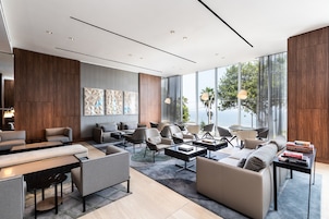 Un moderno hall de hotel con diversas zonas de estar, incluyendo sofás y sillones, organizados alrededor de una mesa de centro. Amplias ventanas ofrecen vistas al exterior.
