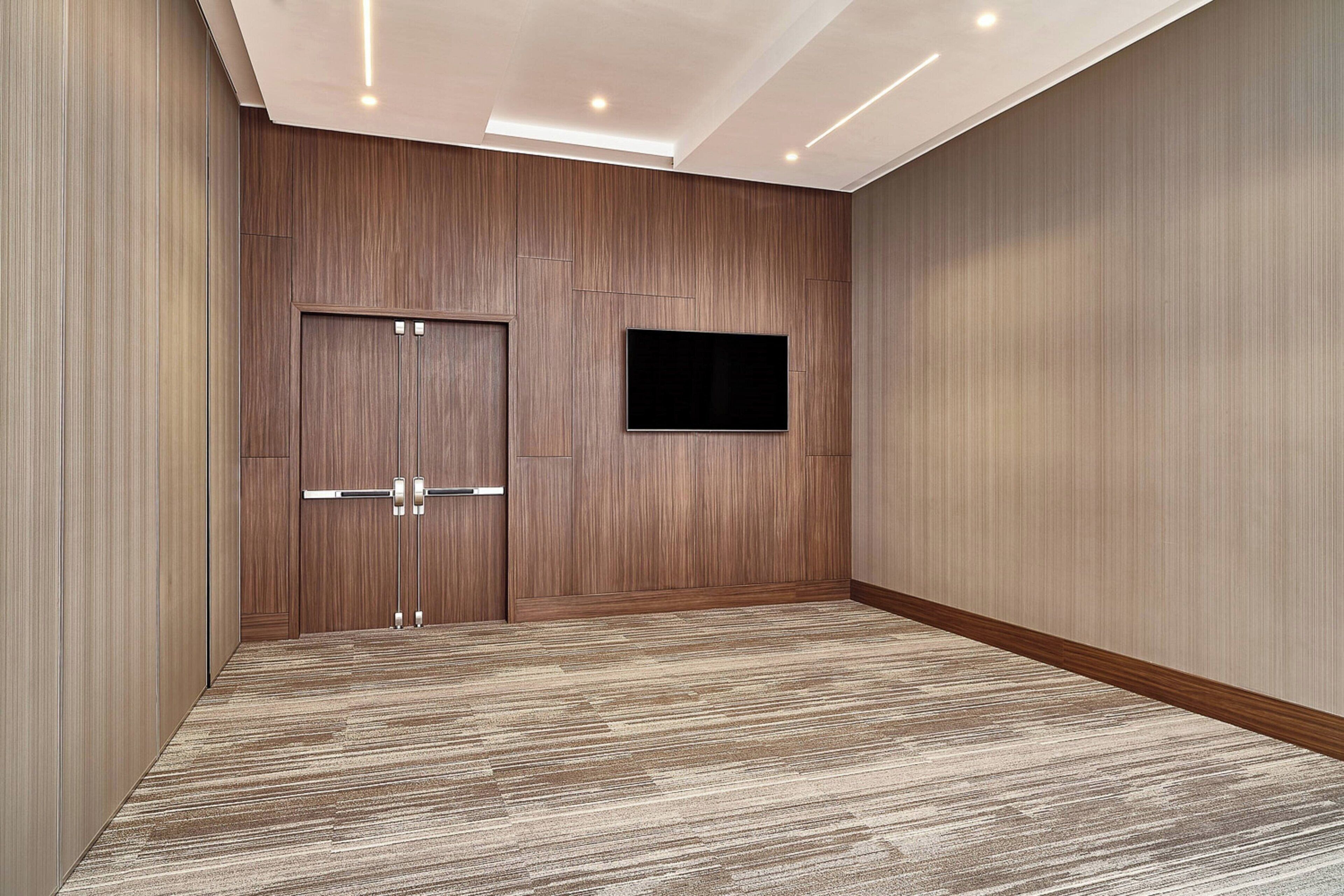 A hotel room with a wooden panel wall, a flat-screen TV mounted on the wall, and a patterned carpet.