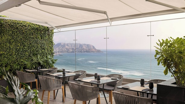Un área de comedor al aire libre con mesas y sillas, vista al océano y una pared verde.