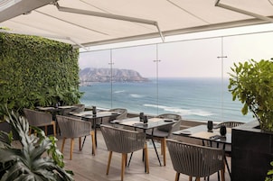 An outdoor dining area with tables and chairs, a view of the ocean, and a green wall.