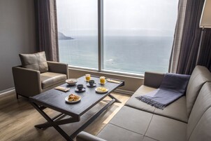 A modern room with a large window overlooking the sea, a dining table set with breakfast, and two sofas.