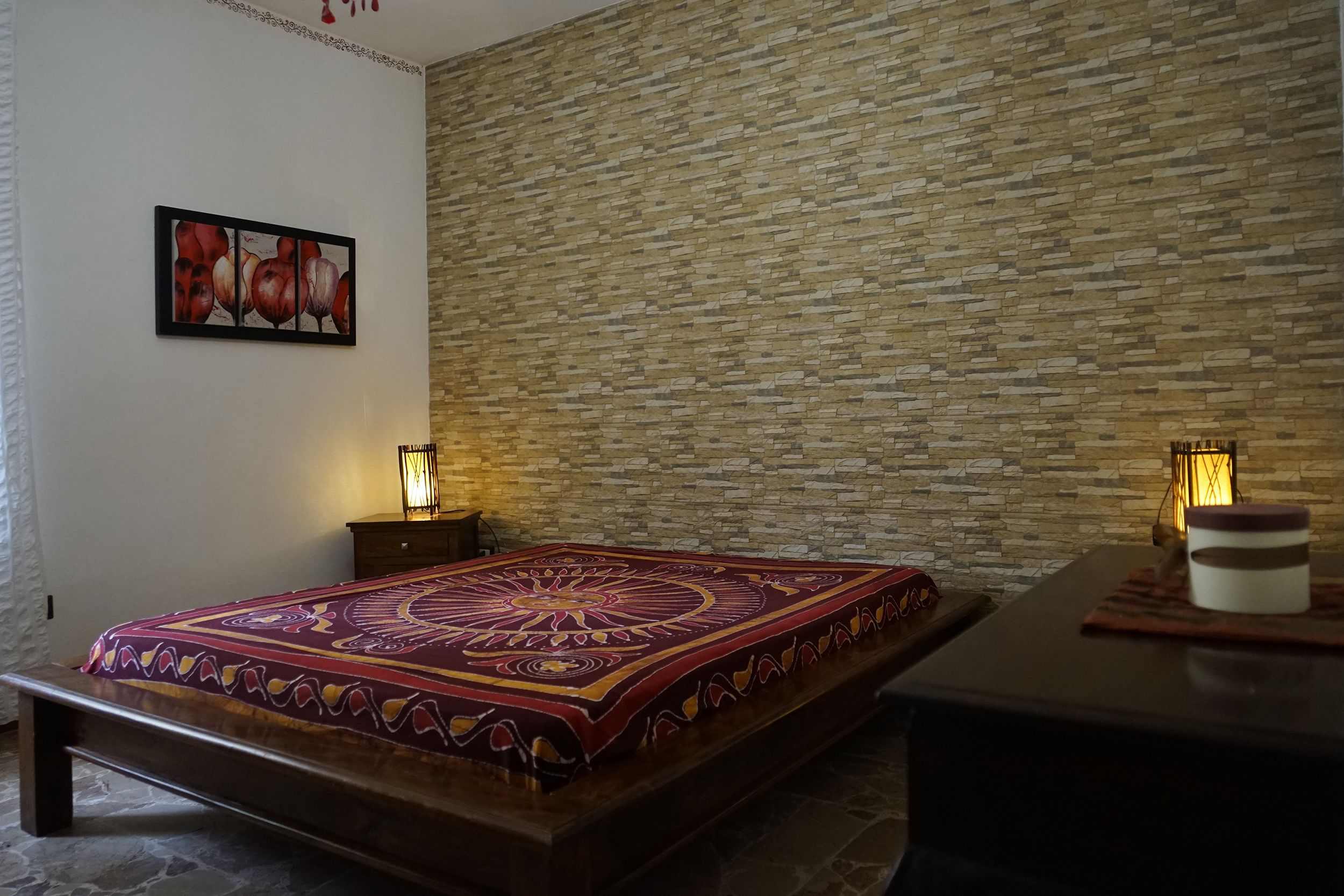 A room with a bed, a nightstand, a table with a candle, and a wall with a patterned design.