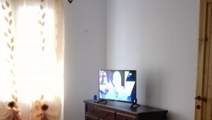 A room with a tiled floor, a television on a wooden cabinet, a window with sheer curtains, and a hanging light fixture. - Casa Barchetta (Alghero)