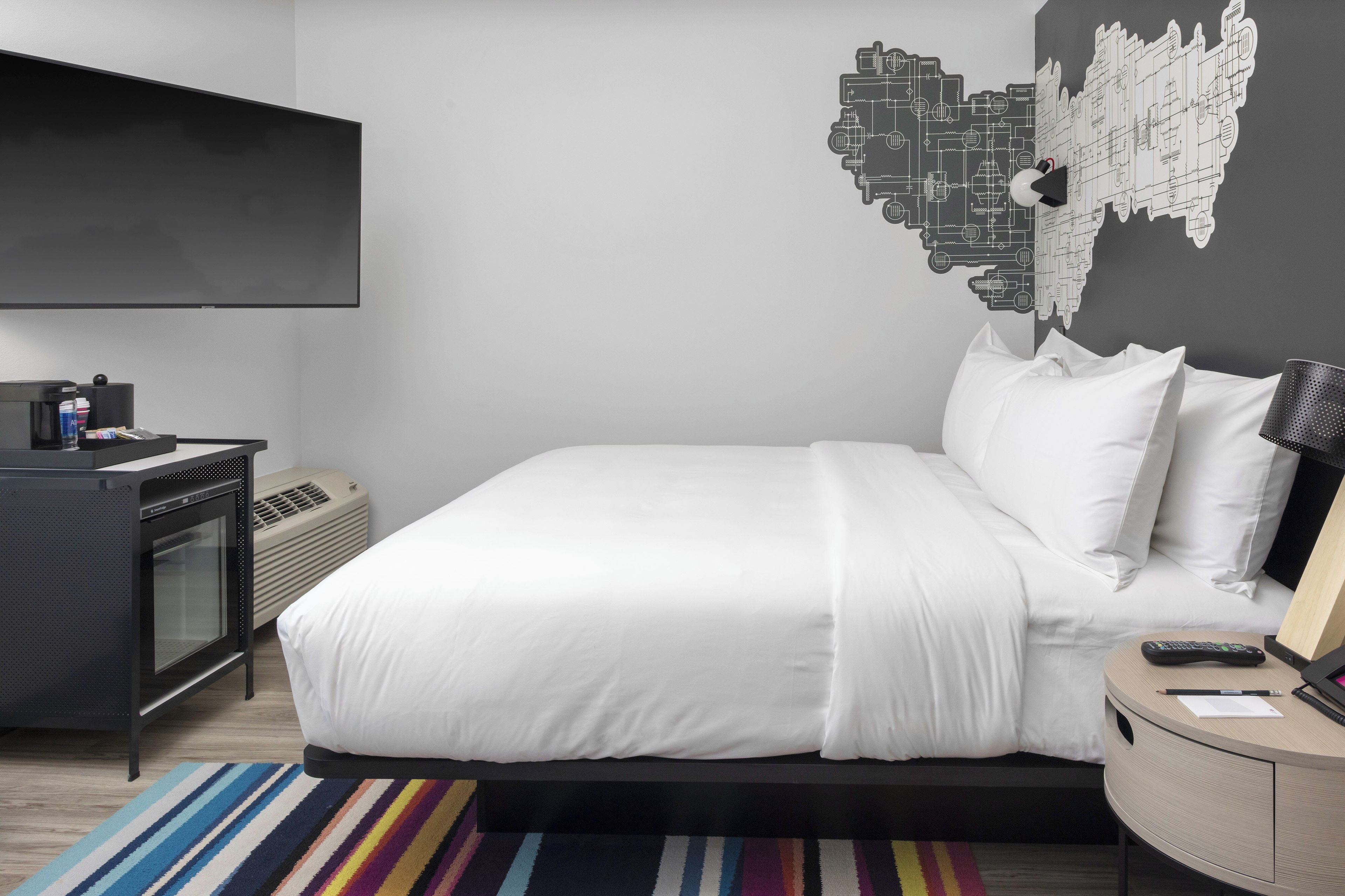A modern hotel room with a large bed, a flat-screen TV, a desk with a computer, and a wall art piece.