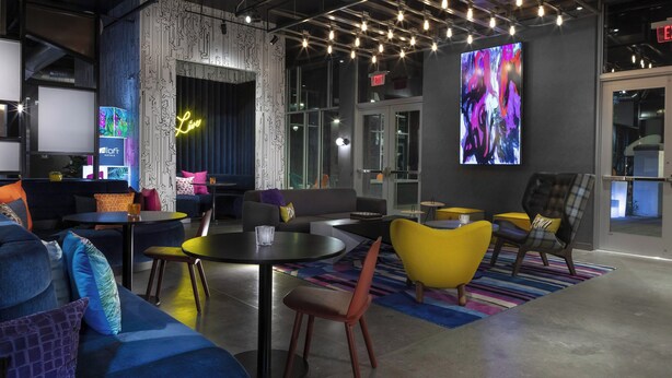A modern lounge area with a mix of seating options including sofas and chairs, a round table, and a large screen displaying abstract art. - Aloft by Marriott Glendale at Westgate (Glendale)