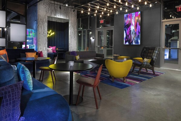 A modern lounge area with a mix of seating options including sofas and chairs, a round table, and a large screen displaying abstract art. - Aloft by Marriott Glendale at Westgate (Glendale)