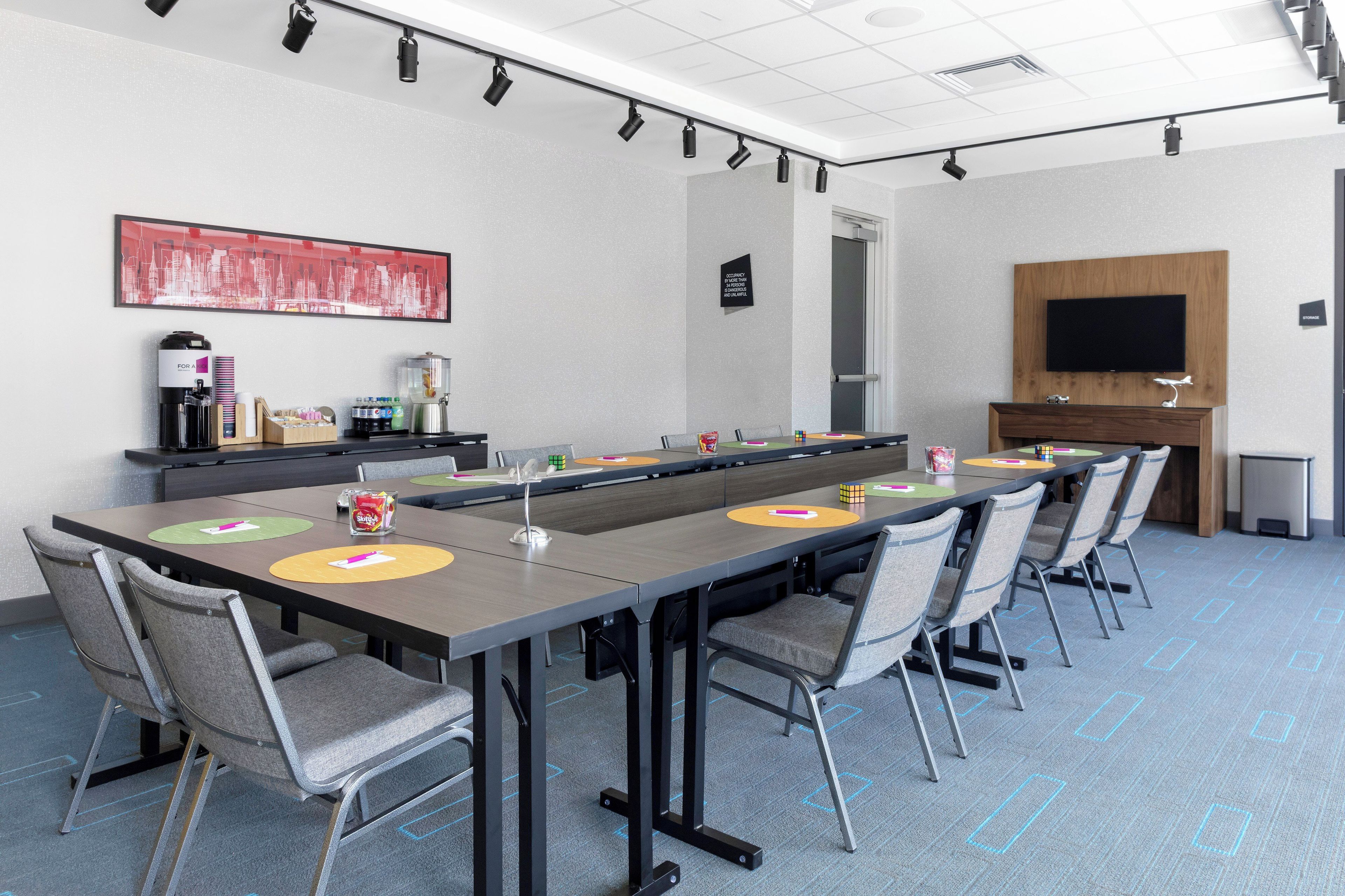 A conference room with a long table, chairs, a coffee station, a painting, a television, and a door.