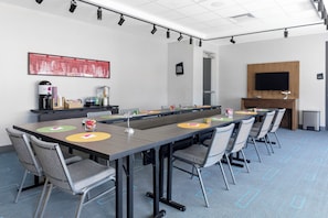 Une salle de conférence avec une longue table, des chaises, un coin café, un tableau, un téléviseur et une porte.