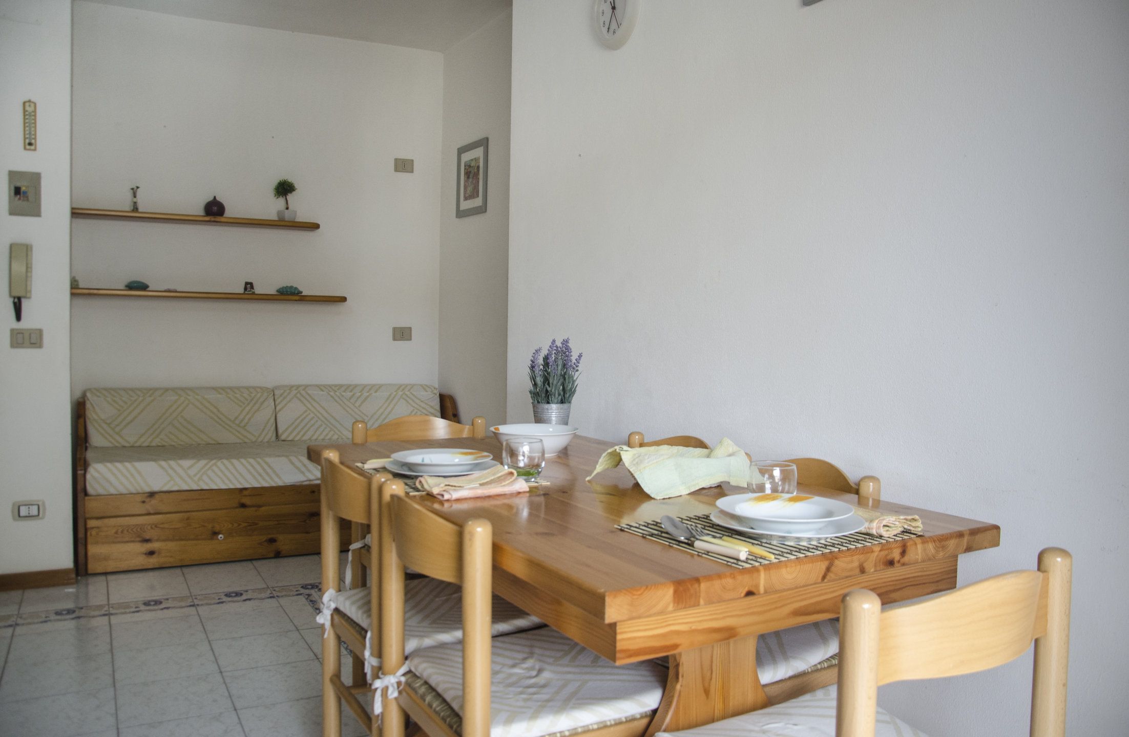 Un comedor con una mesa de madera dispuesta para dos personas, sillas, un sofá y un estante pequeño con objetos decorativos.