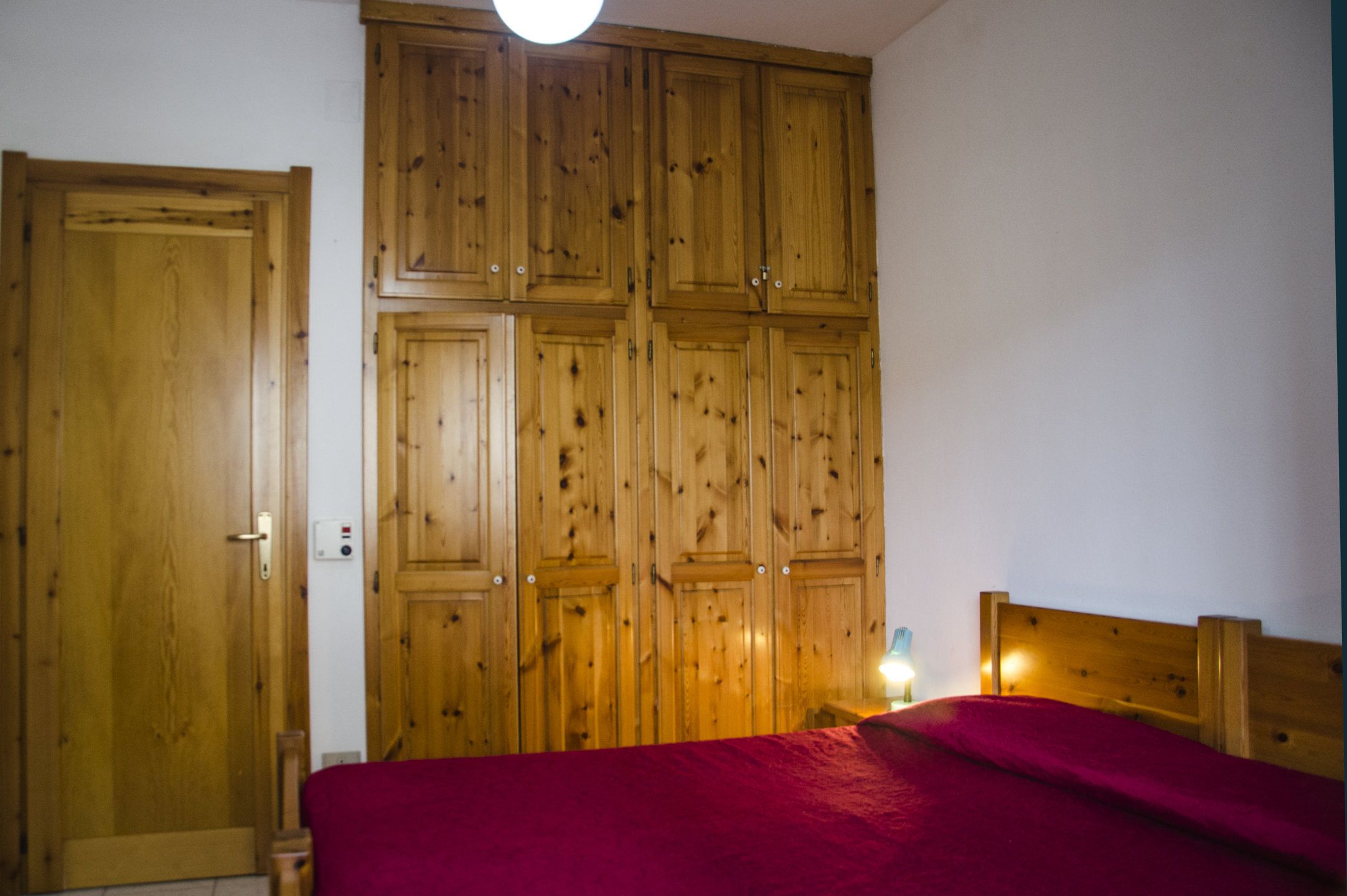 A bedroom with a wooden wardrobe, a bed with a red blanket, and a nightstand with a lamp.
