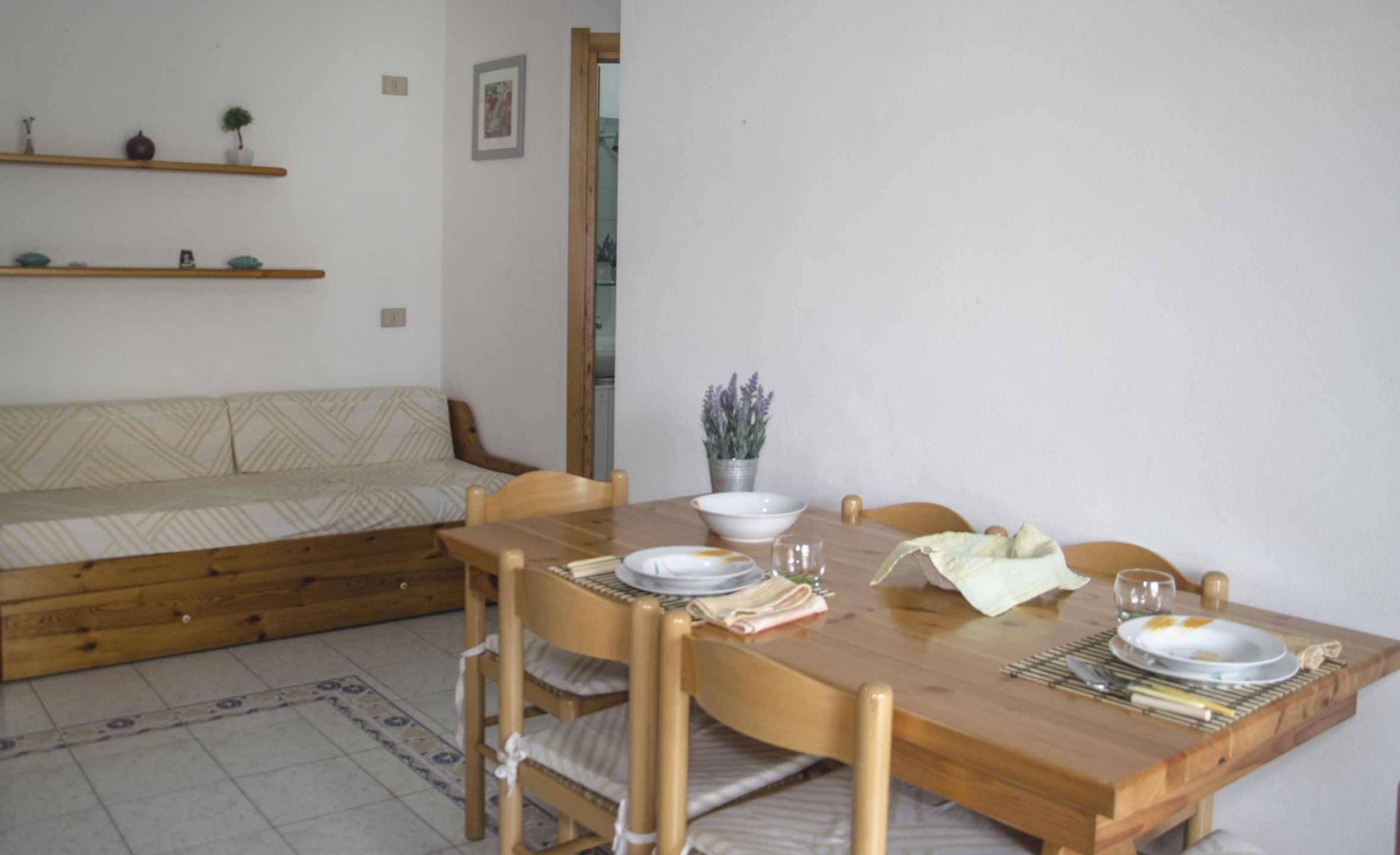 Zona pranzo con tavolo in legno apparecchiato per quattro persone, divano e una piccola pianta in vaso su una mensola.