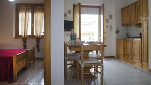 A room with a bed, a dining table with chairs, a kitchen area with wooden cabinets, and a window with curtains. - Casa Mostella (Alghero)