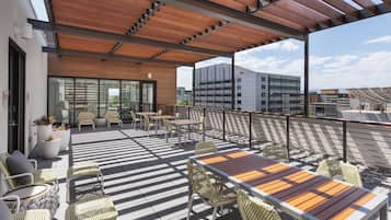 An outdoor terrace with wooden tables and chairs, a pergola, and a view of buildings in the background.