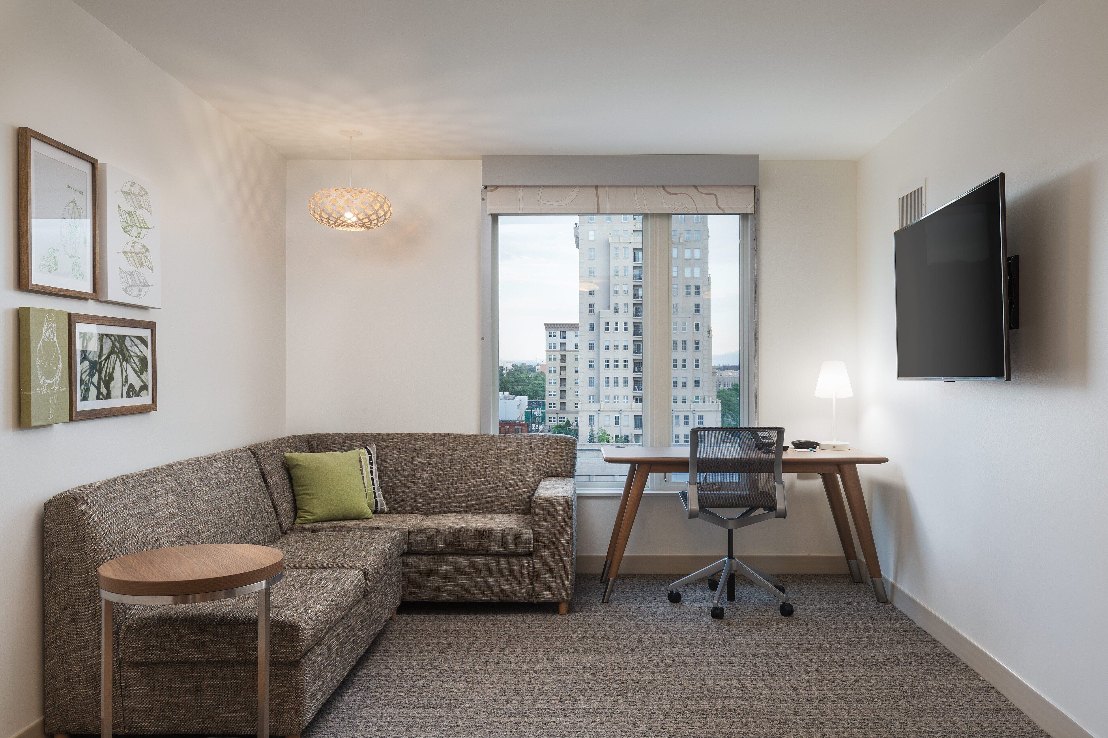 A modern living room with a sofa, a small round table, a desk with a chair, a TV mounted on the wall, and a large window with a view of high-rise buildings.