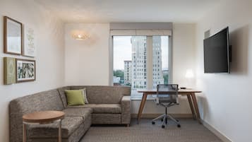 A modern living room with a sofa, a small round table, a desk with a chair, a TV mounted on the wall, and a large window with a view of high-rise buildings.